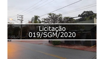 Fotografia que mostra rua, casas e na frente das casas tem árvores e no fim da rua tem poste de luz, o céu está nublado, no meio tem faixa transparente escrito Licitação 019/SGM/2020