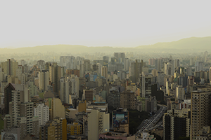 Arte com uma foto aérea da cidade de São Paulo no fundo sobreposta com ícones que fazem referência ao coronavírus.