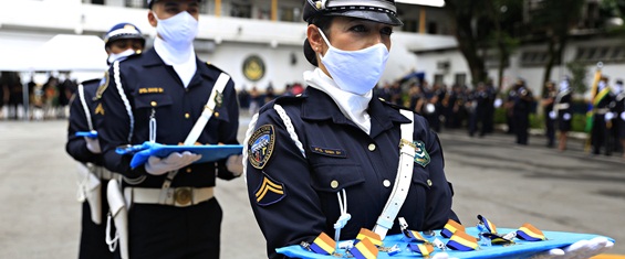 #PraTodosVerem- Tres GCMs de uniforme azul, mascara branca, carregam em uma bandeja azul , varias medalhas com fitas, nas cores azul, branco e amarelo. Ao fundo imagem desfocada de pessoas.