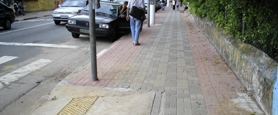 Foto mostra pessoa andando em calçada acessível, com rampa de acessibilidade em primeiro plano.