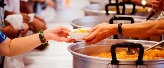 #PraTodosVerem Grandes panelas em refeitório cheias de comida, tendo como primeiro plano uma pessoa com um prato (só aparece o braço e a mão) sendo servida.