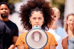 Foto de uma mulher negra, vestindo camiseta laranja e segurando um megafone em frente da boca.