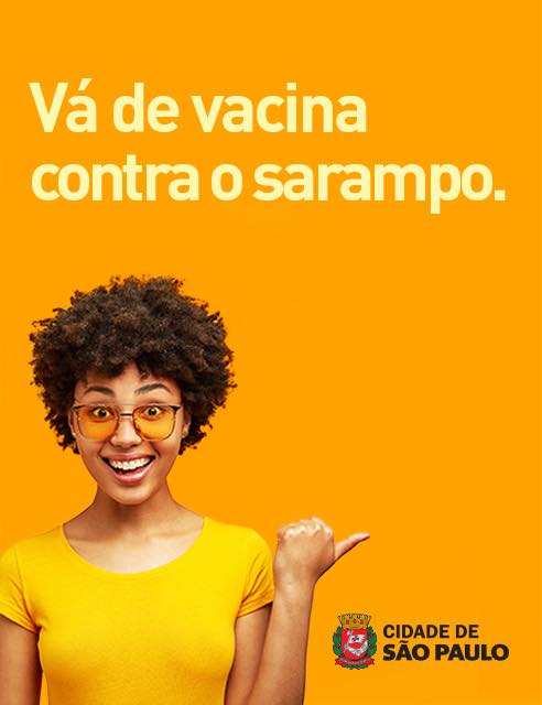 Foto de uma jovem vestida de amarelo e chamando, com o polegar, as pessoas para se vacinarem.