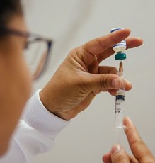 Foto de uma enfermeira de óculo com uma ampola da vacina na mão preparando a seringa para aplicação.