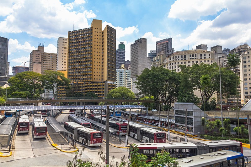 Fotografia que mostra um Terminal de ônibus, com vários ônibus e no fundo da imagem com vários prédios.