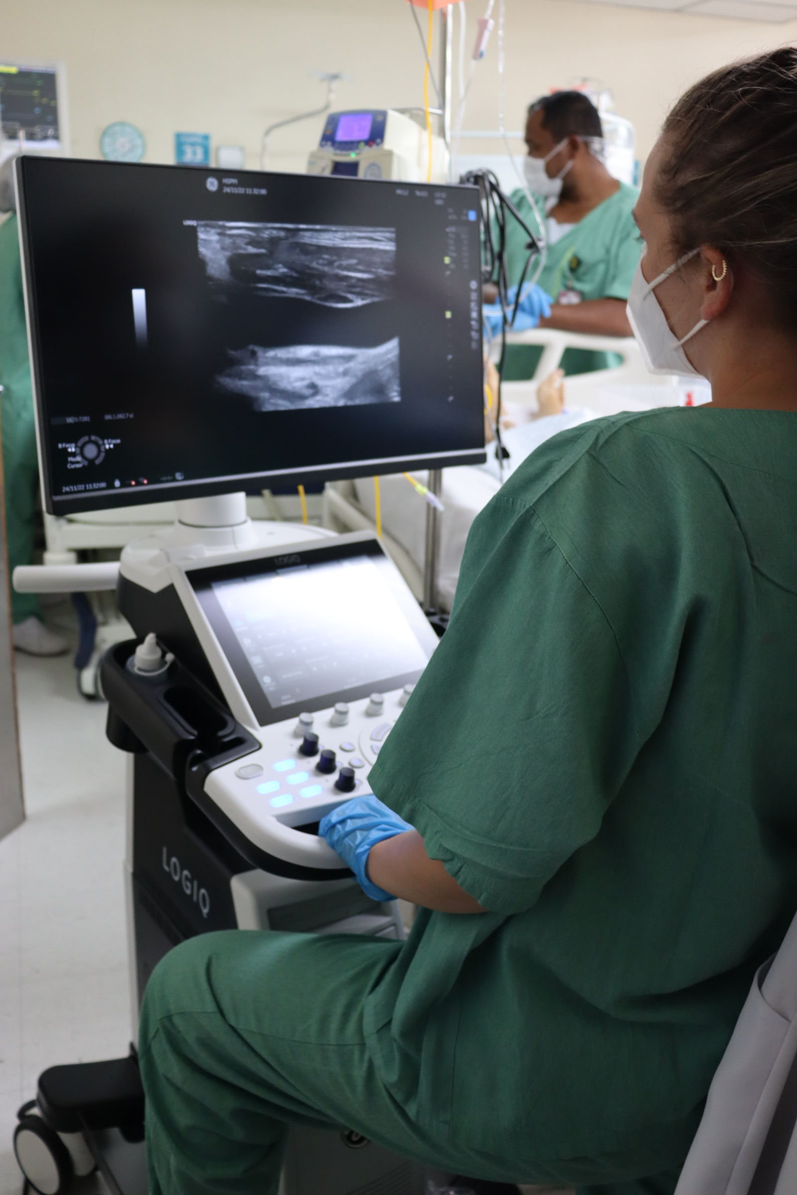 Foto mostra um quarto de hospital e uma enfermeira sentada à frente do novo equipamento de ultrassom, utilizando o equipamento.