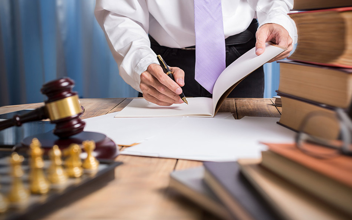 No centro da foto, uma pessoa curvada sob uma mesa assina documentos. Ao seu redor, na mesa de madeira, encontram-se um martelo de decisões judiciais e livros empilhados. 