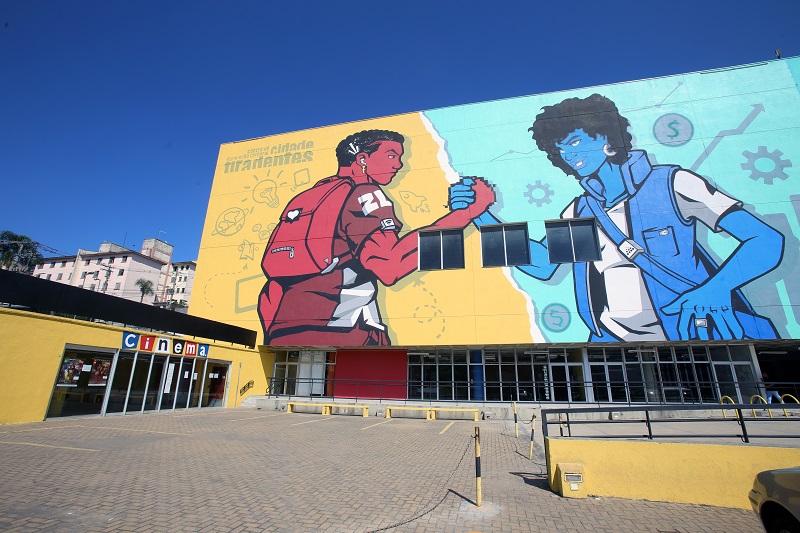 foto da entrada de cinema a esquerda, ao lado prédio com fachada pintada com dois persoangens cobrindo toda a fachada e dando as mãos, a esquerda um homem na cor vermelha e a direita uma mulher na cor azul.