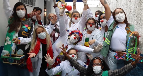 Na imagem há várias pessoas vestindo jaleco branco do SUS, máscara de proteção e estão posando para a foto. Algumas pessoas estão com os braço levantados e usam nariz de palhaço e rosto pintado.