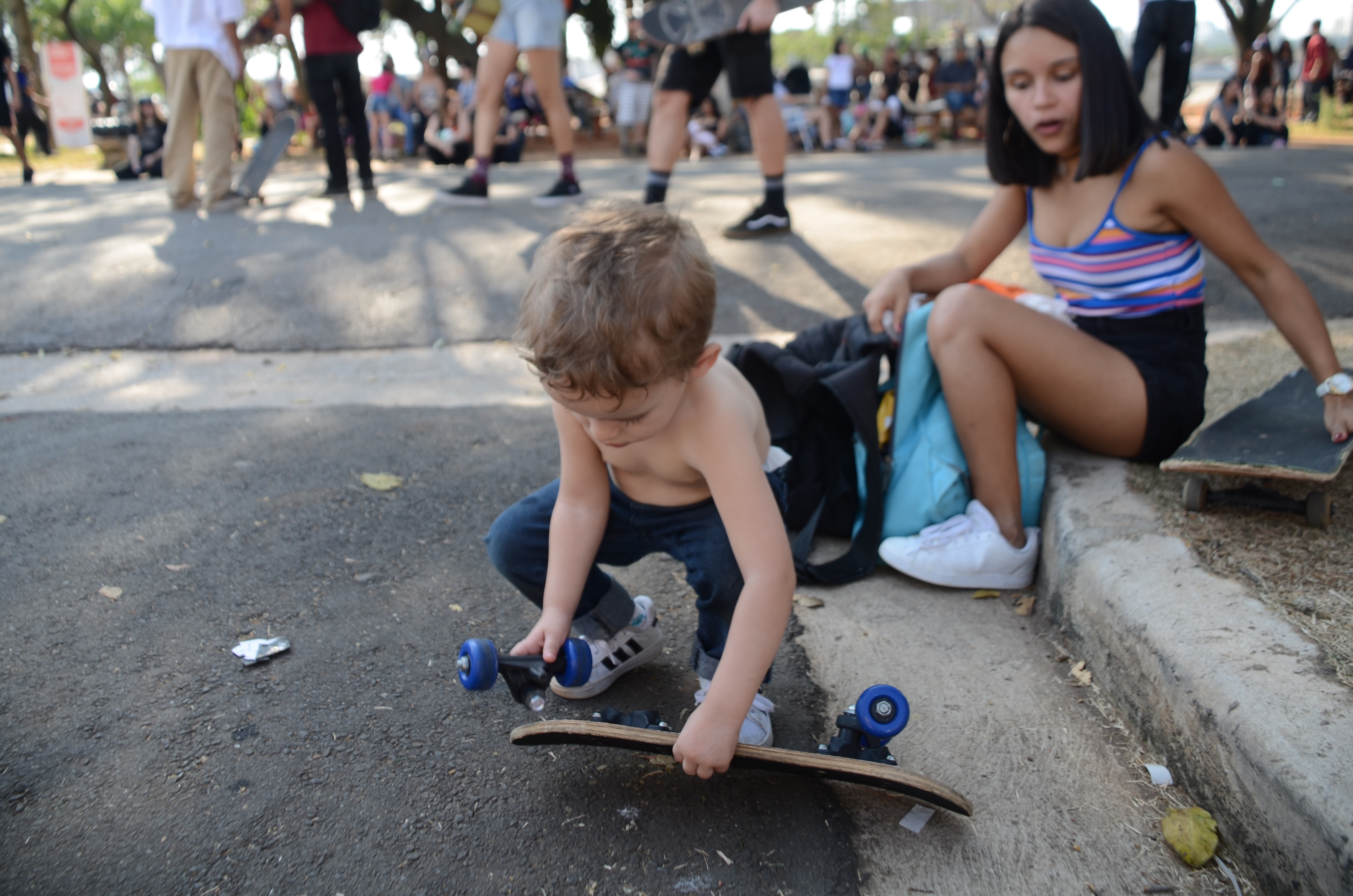 Criança brinca com peças de um skate, agachada no asfalto.