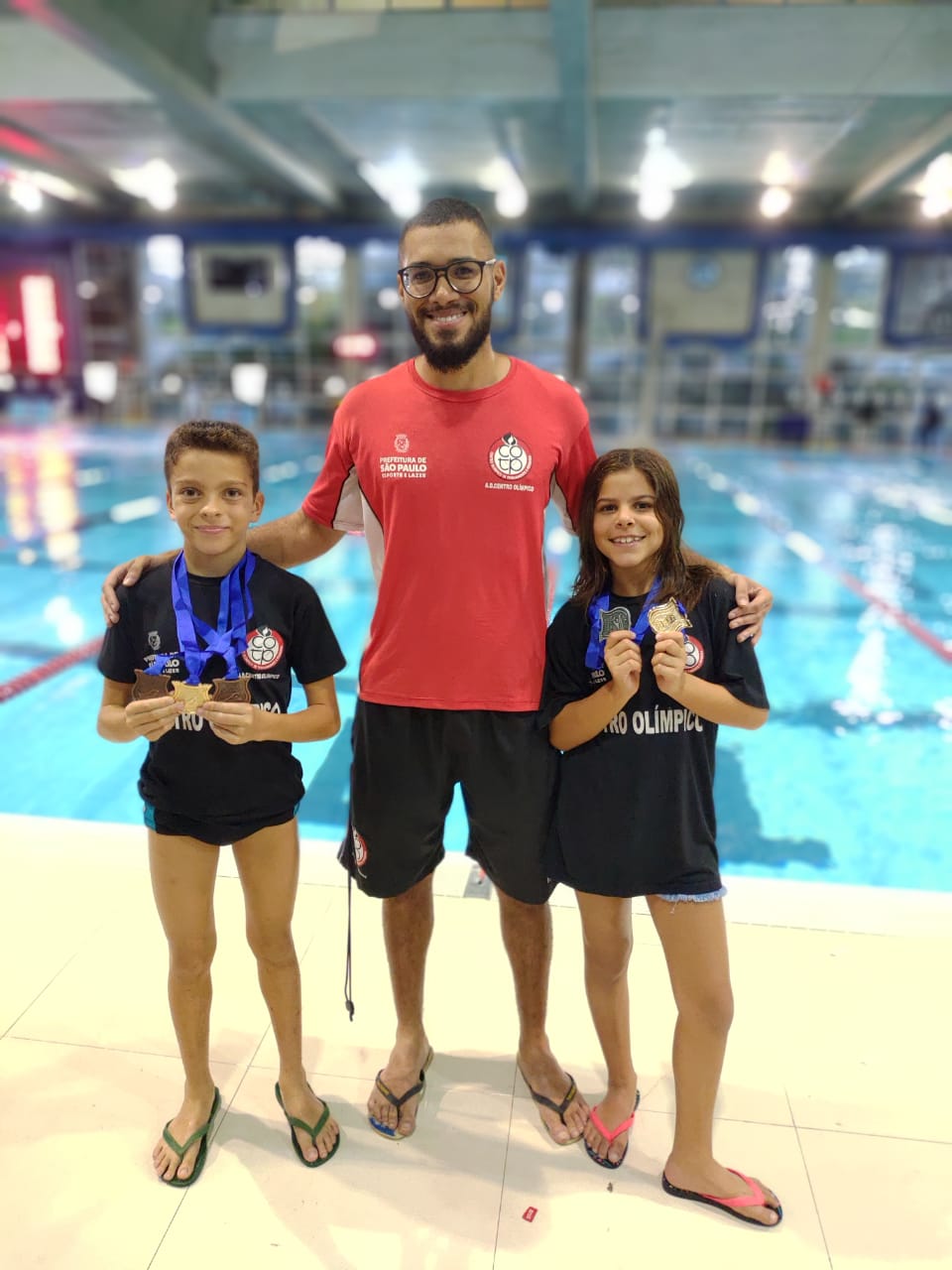 Dois atletas posicionados segurando suas medalhas com o técnico no meio na frente da piscina