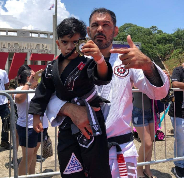 Lutador de MMA Minotauro segura no colo uma criança que exibe medalha pendurada no pescoço.