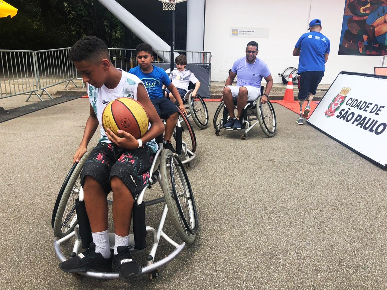 A imagem mostra quatro pessoas em cadeiras de rodas. A primeira da fila está com uma bola de basquete no colo.