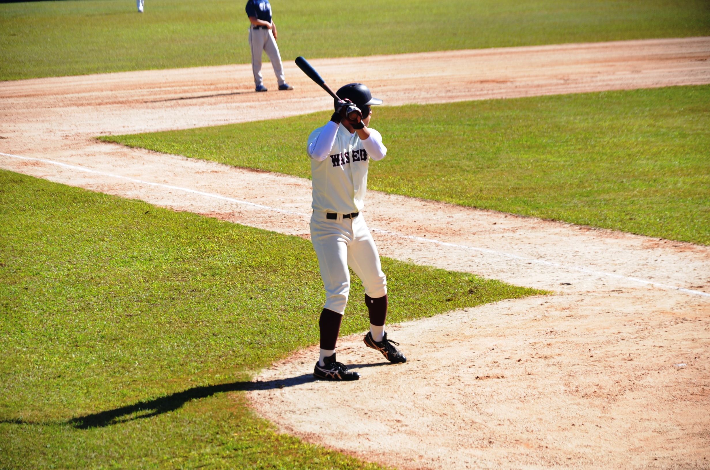 Na imagem um jogador de beisebol com equipamento e taco nas mãos pronto para rebater a bola, em um campo deste esporte, em um dia ensolarado.