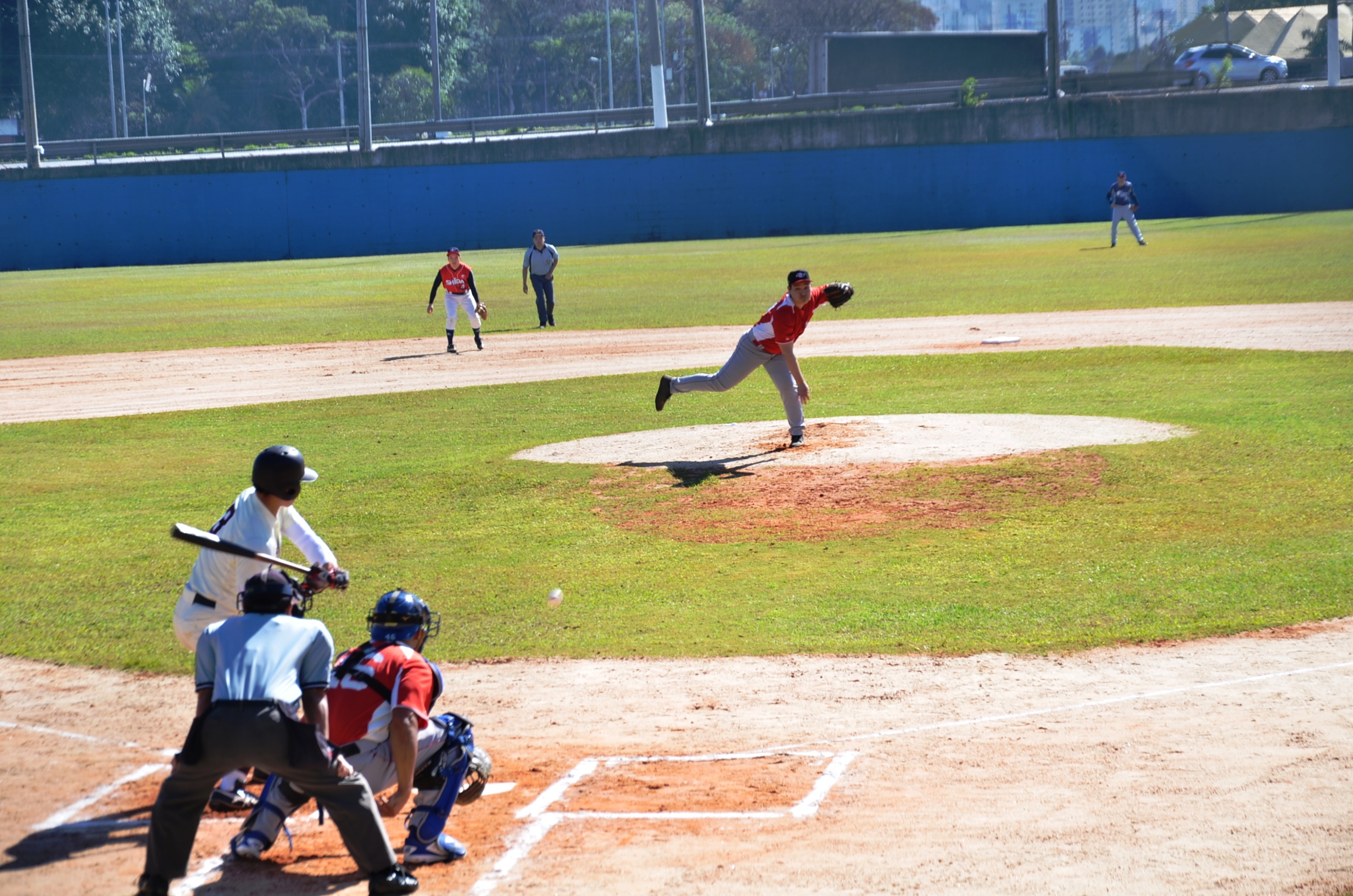 Na imagem jogadores de beisebol com equipamentos, jogando a modalidade em um dia ensolarado.