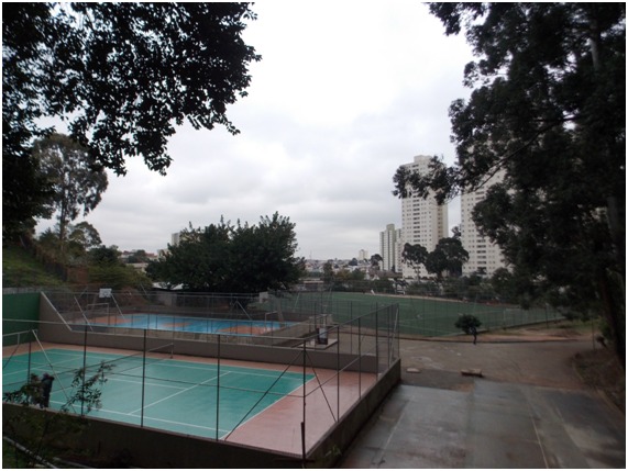 Na imagem um Centro Esportivo com árvores, duas quadras poliesportivas, um campo de futebol e no fundo prédios em um dia nublado.