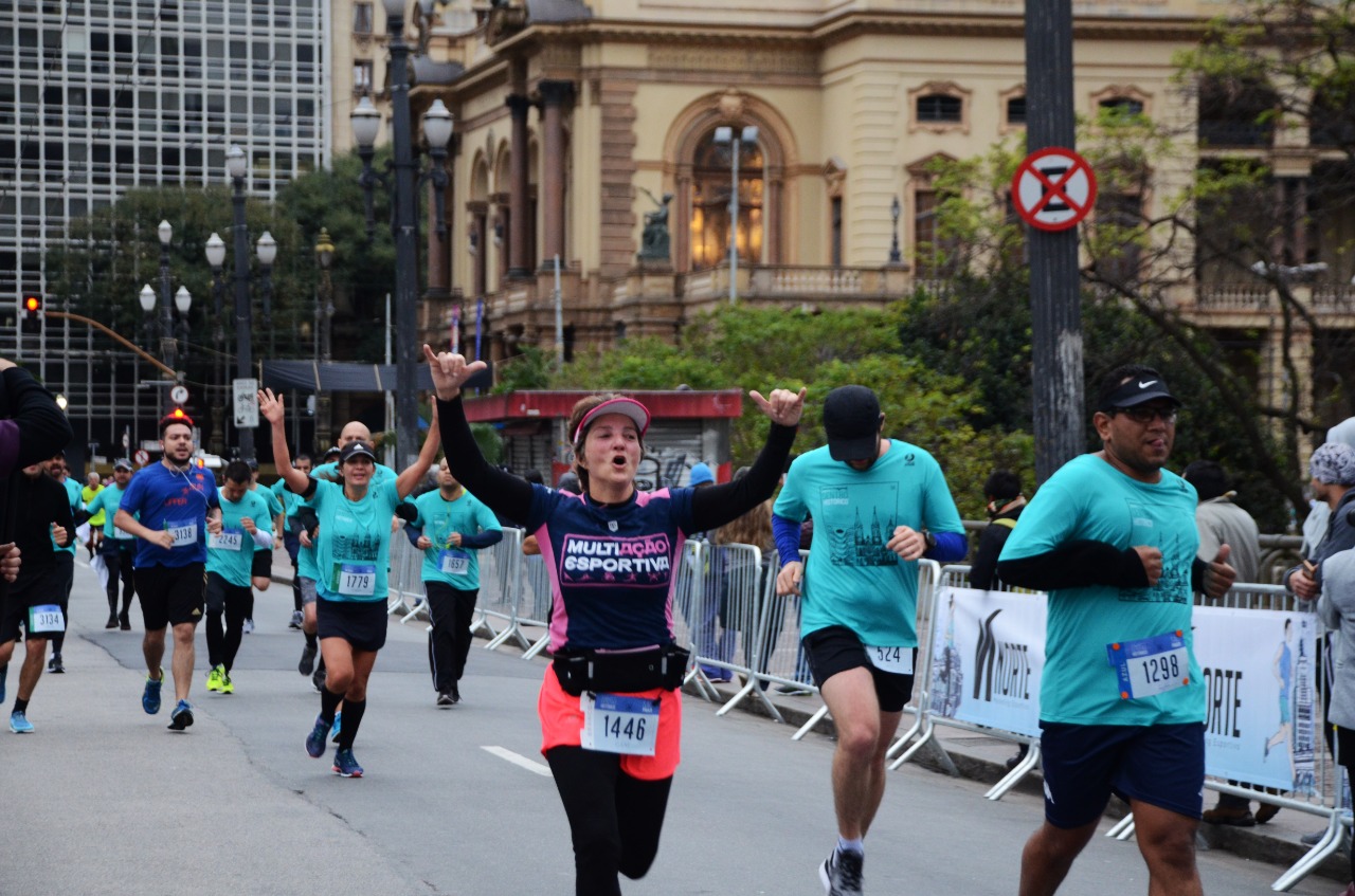 Foto de diversas pessoas em uma corrida de rua. Em destaque, uma mulher ergue os braços em celebração. Ao fundo, é possível ver o Teatro Municipal de São Paulo.