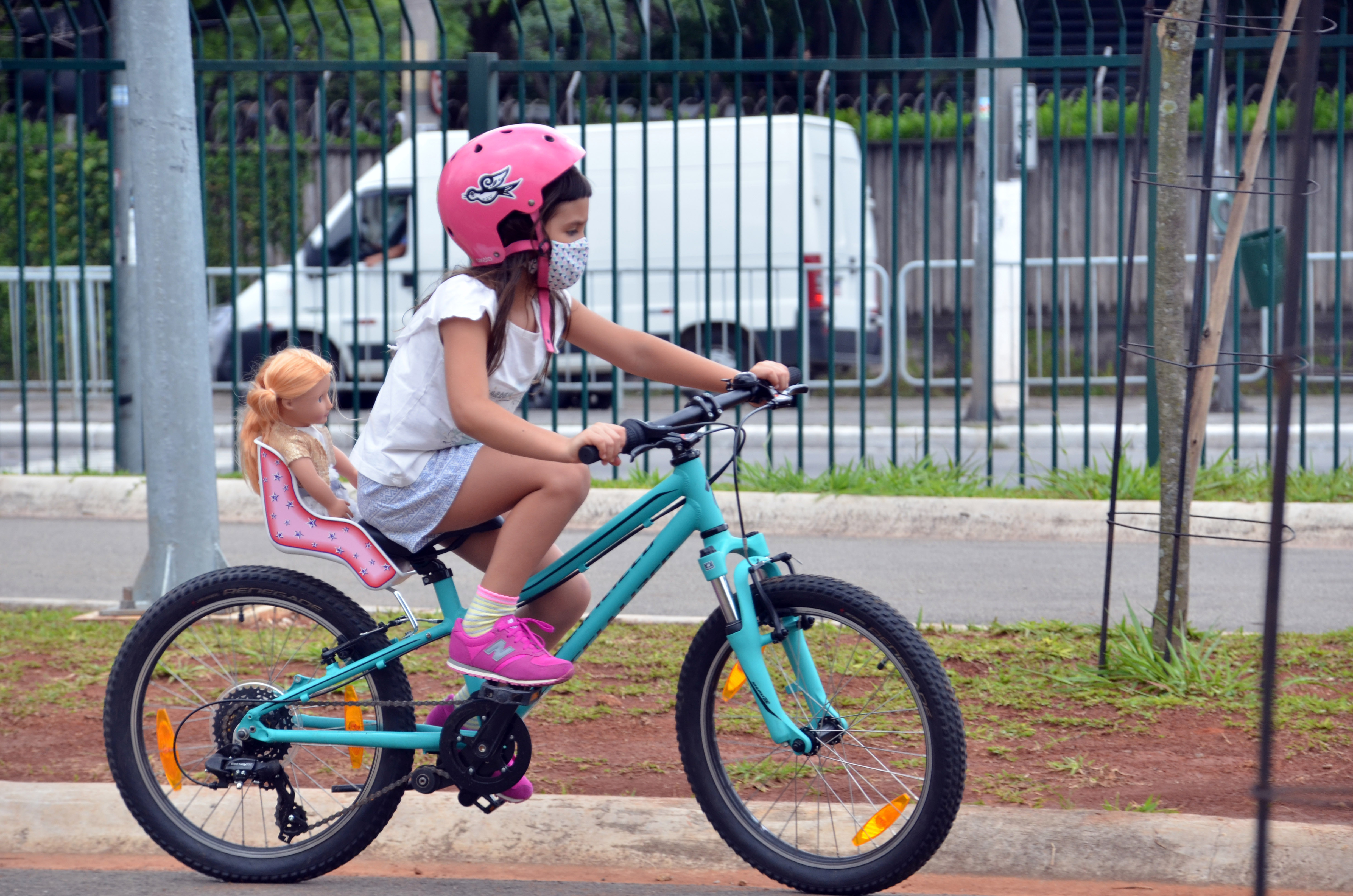 Menina anda de bicicleta no Parque das Bicicletas usando máscara. Na cadeira de trás da bicicleta, a menina leva uma boneca.