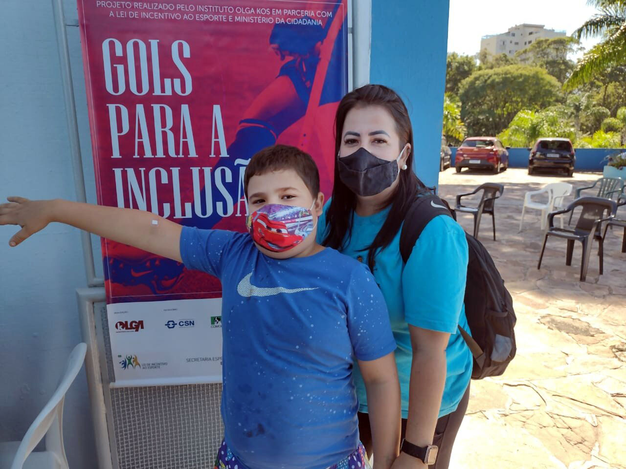 Mãe e filho inscritos no projeto Gols para a Inclusão posam para foto em frente ao banner do programa.