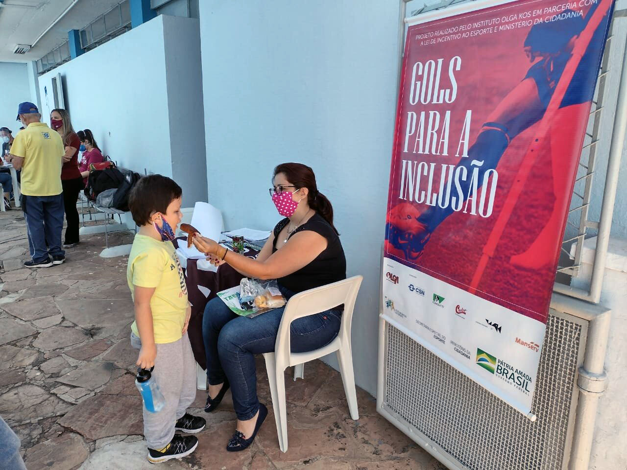 Mãe dá bolinho ao seu filho enquanto aguarda finalização do processo de inscrição no Centro Esportivo Pirituba. Banner o "Gols para Inclusão" aparece à direita.