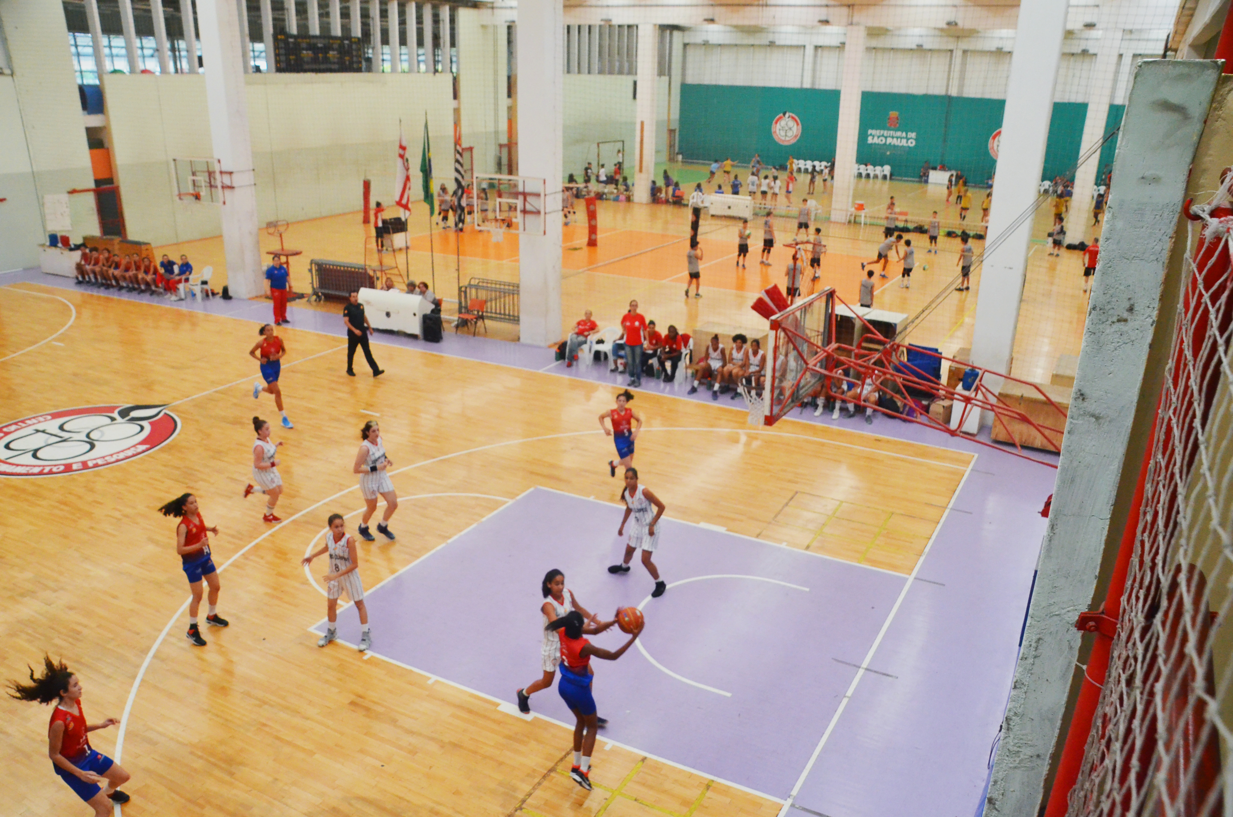 Imagem da visão de cima das quadras do Centro Olímpico, com a de basquete em primeiro plano, seguida pelas de vôlei e handebol. Há atletas jogando.