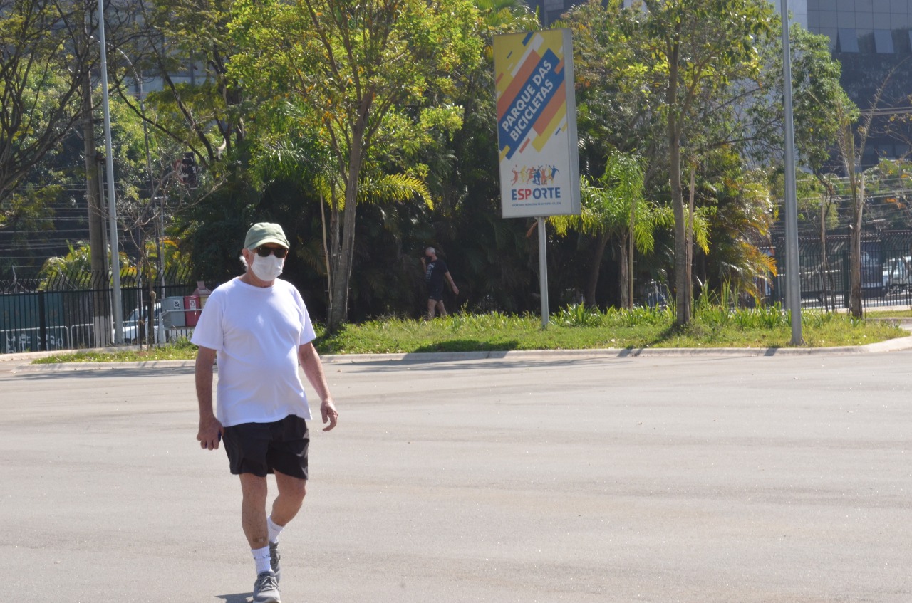 Na imagem, homem de boné, óculos de sol e máscara caminhando no Parque das Bicicletas em um dia ensolarado. Atrás árvores e a placa com a escrita 