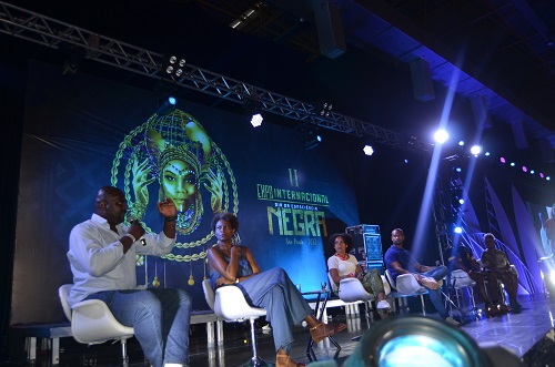 Na imagem, debatedores do Racismo no Esporte sentados em cadeiras em cima do palco.