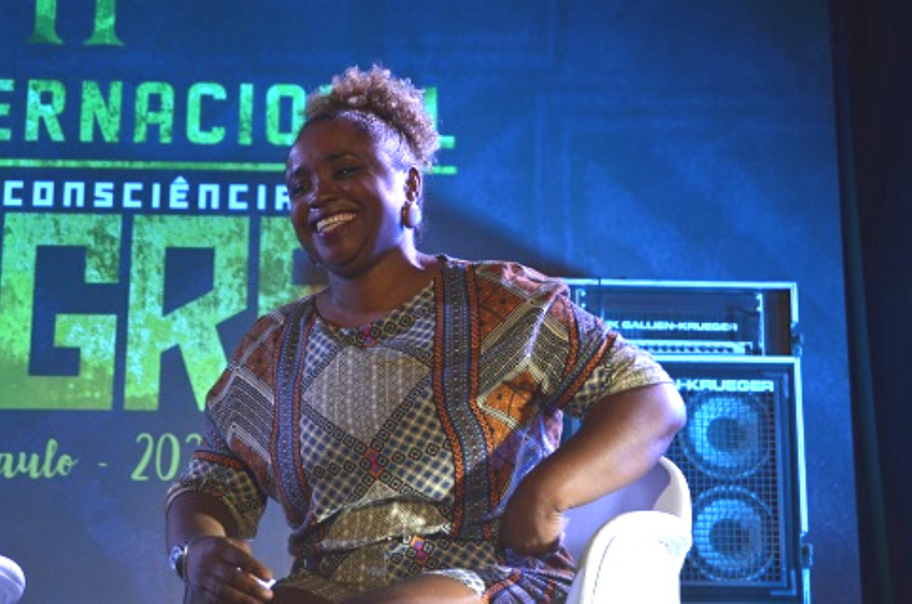 Na imagem, Daiane dos Santos está sorrindo sentada, com o fundo da II Expo Internacional Consciência Negra