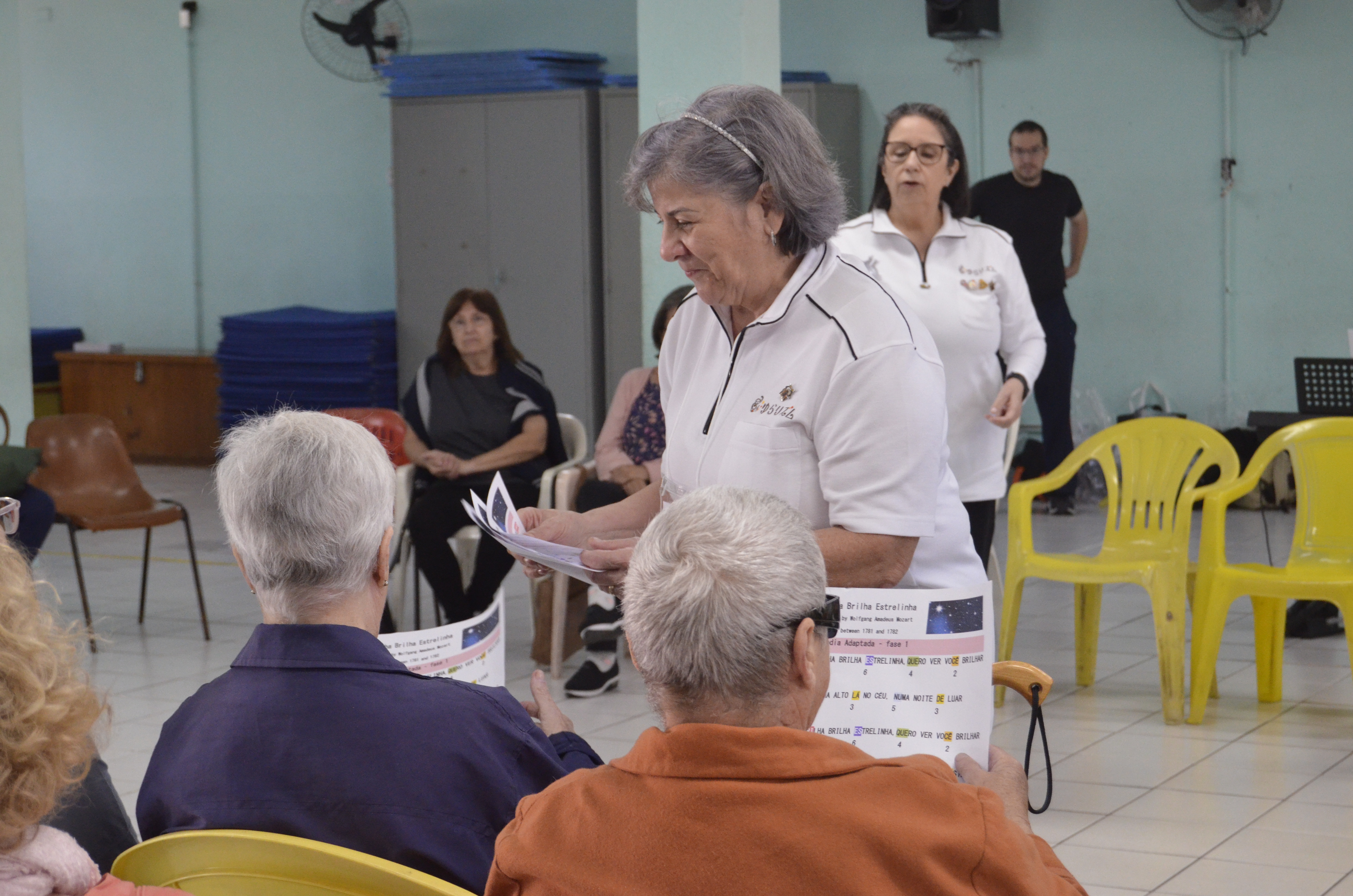 Na imagem, munícipes sentados, recebendo partituras das organizadoras.