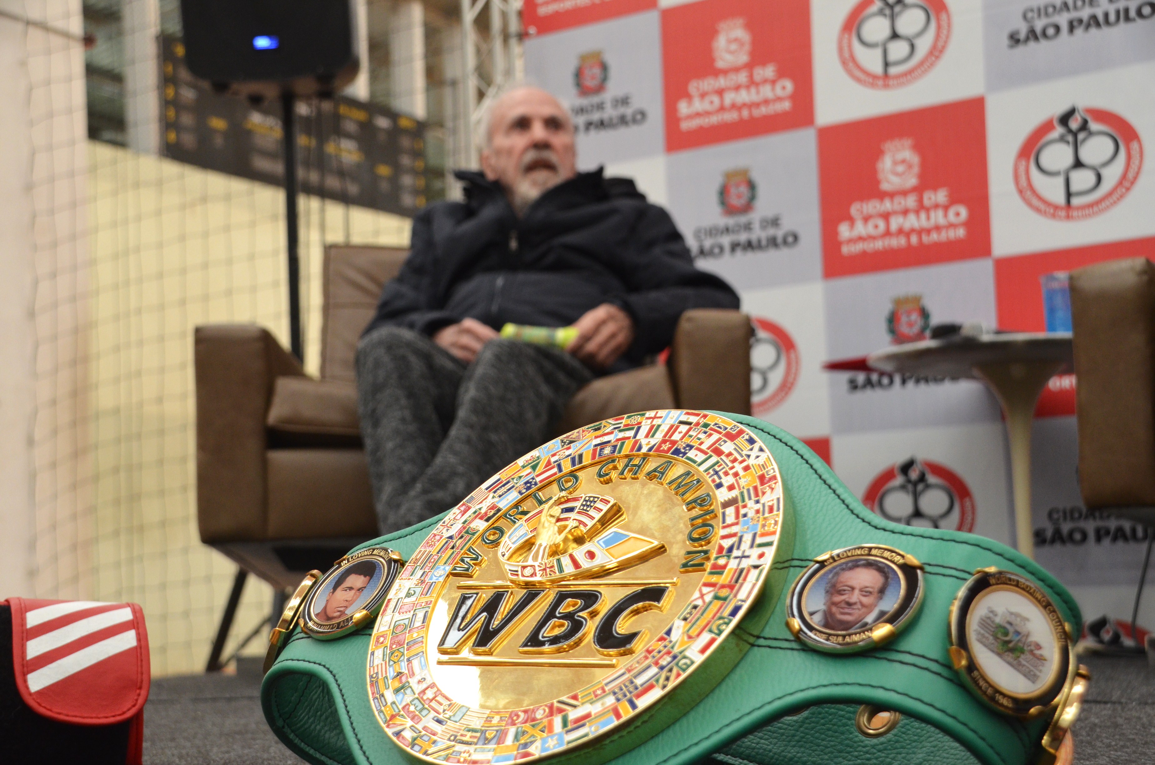 Foto do boxeador Eder Jofre sentado em um sofá com seu cinturão numa mesa a frente.