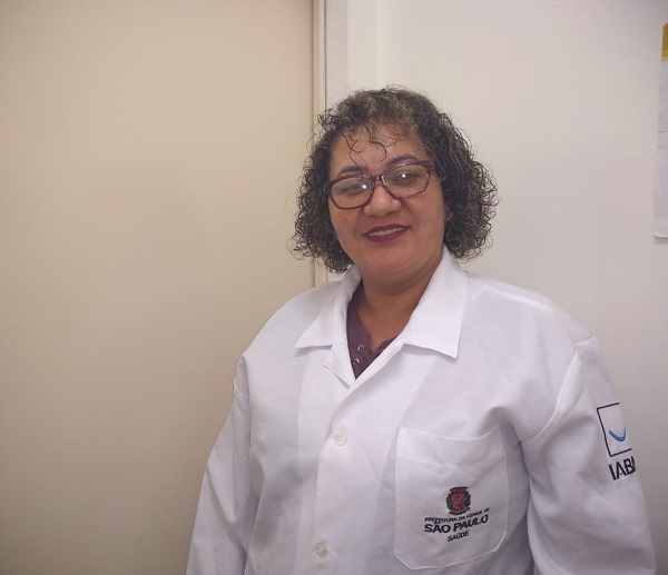 #PraCegoVer: Enfermeira vestindo um jaleco branco e óculos de grau. Sorrindo para foto.