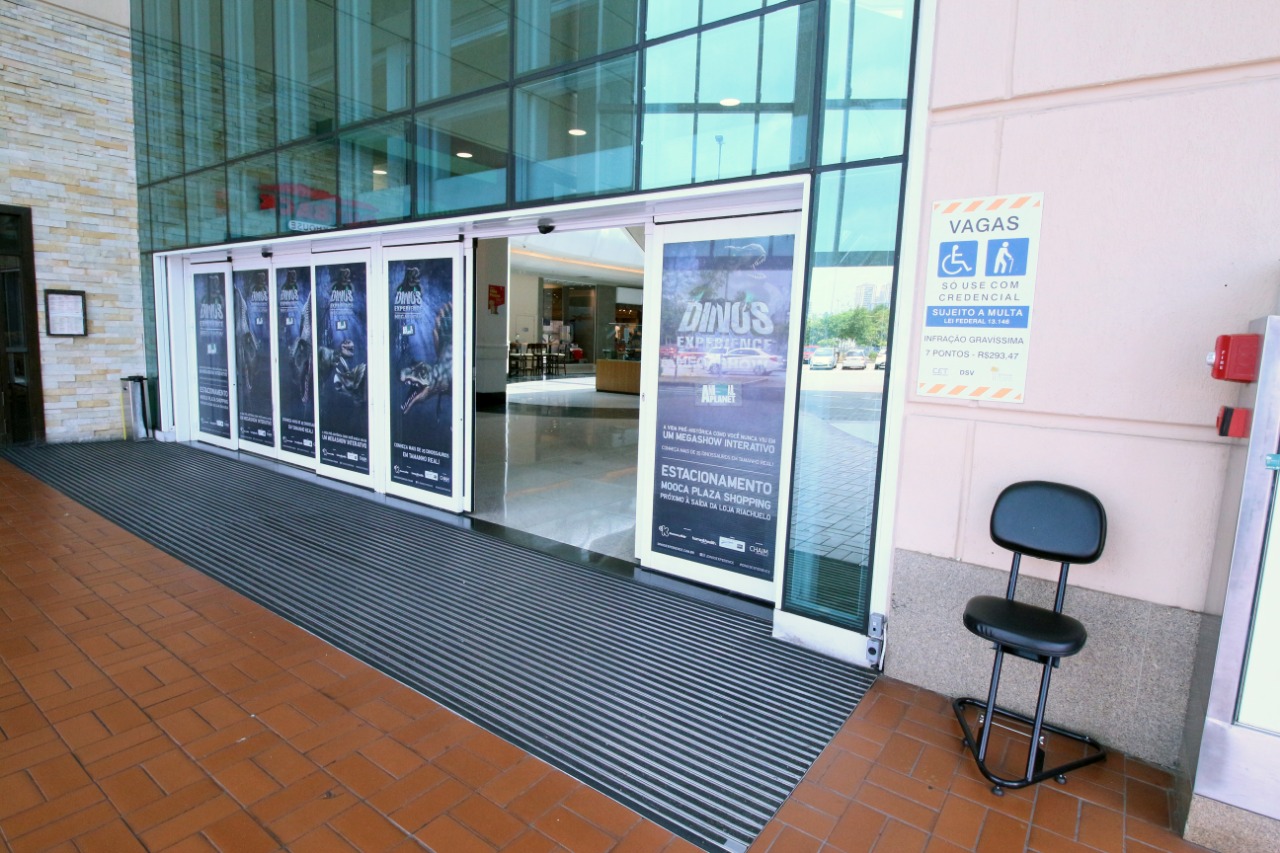 Entrada do shopping, com portas de vidros e automáticas, com um comunicado sobre vagas reservadas. 