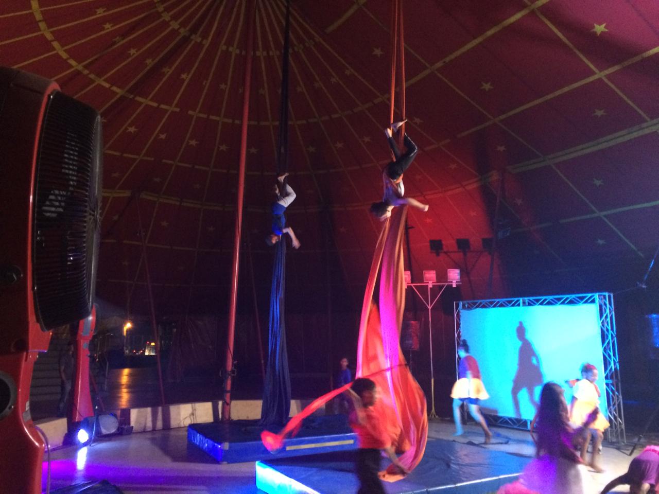 Dois trapezistas fazendo performances. Um com pano azul e outro com pano vermelho em picadeiro do Circo Escola. Ao fundo duas pessoas em pé com telão branco atrás de si. 