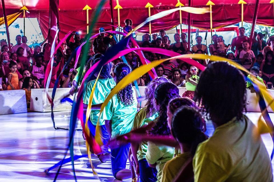 Oito crianças vistas de costas fazem movimento circular com fitas coloridas em picadeiro do Circo Escola.