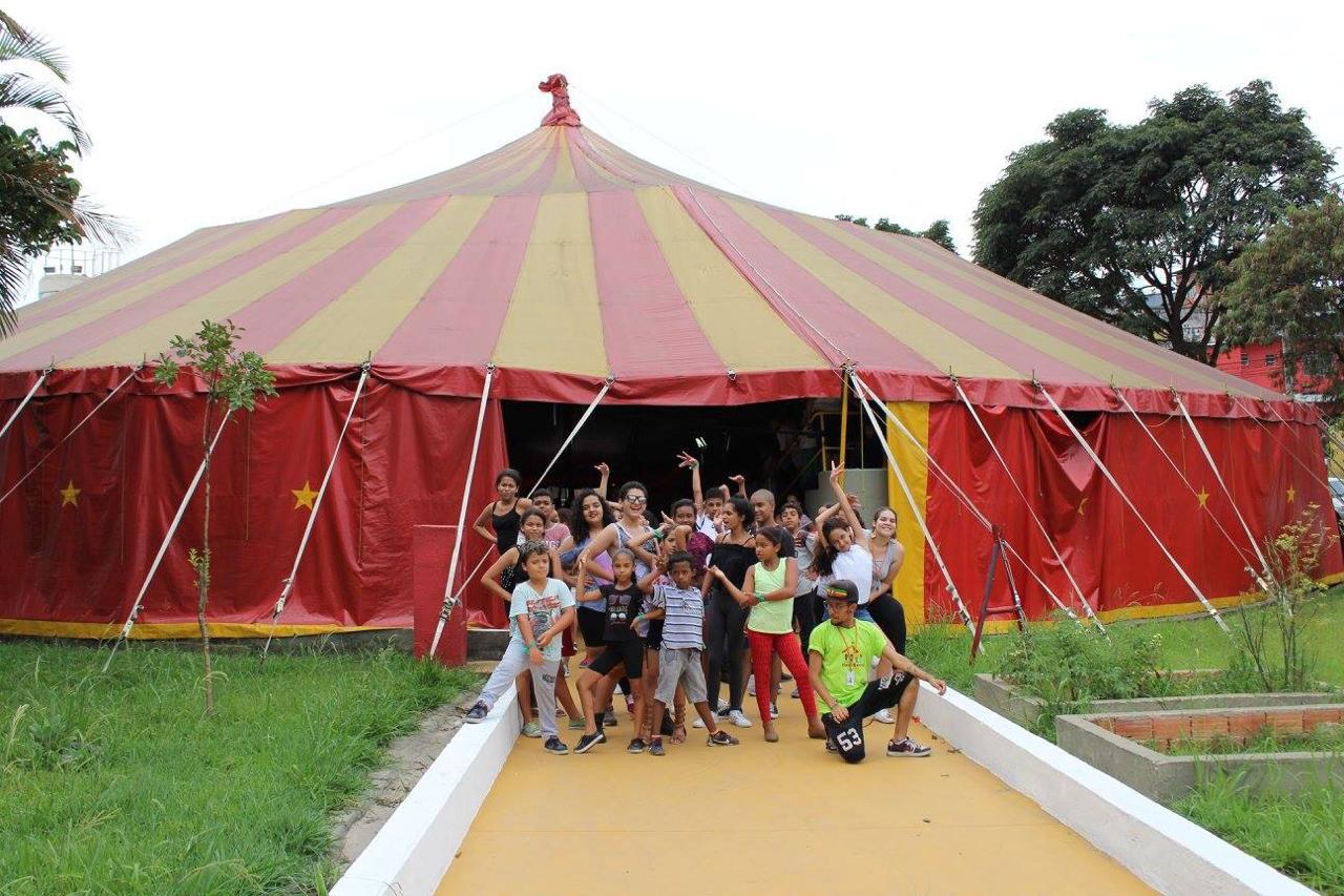  Conviventes do Circo Escola reunidos na entrada de lona vermelha em pose para foto. Alguns estão com os braços levantados e outros agachados à frente.