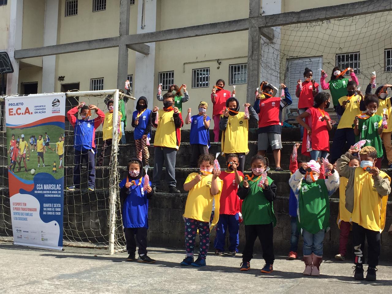 Crianças participantes da atividade, usando coletes que separam as equipes, posam para foto na quadra após receberem as medalhas de participação.
