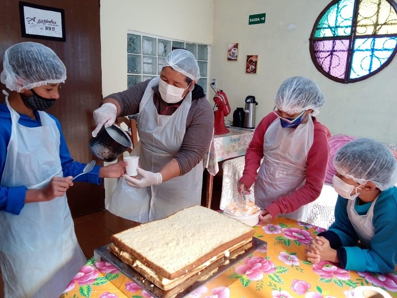 A oficineira Edna Lima ajuda três crianças durante a decoração do bolo.