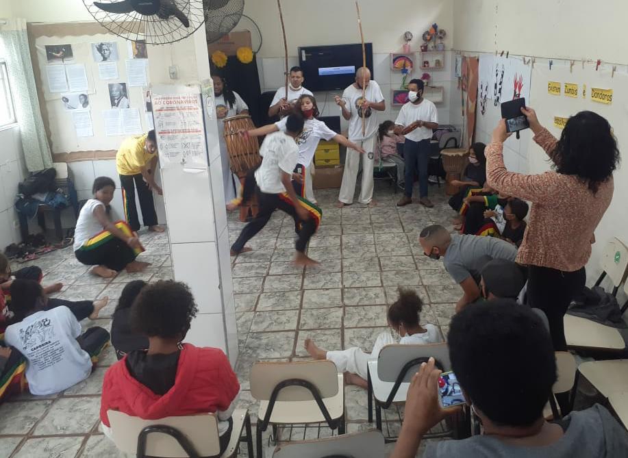 Quatro crianças realizam a atividade de capoeira enquanto seus orientadores tocam os instrumentos de percussão. Outras crianças assistem e uma mulher grava a apresentação com o celular.