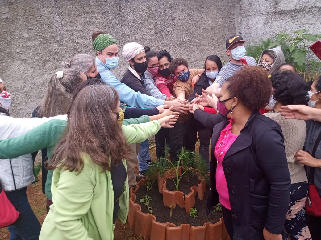 Diversos conviventes do serviço colocam suas mãos umas sobre as outras, simbolizando a união, em volta de uma das plantações feitas na horta do local.