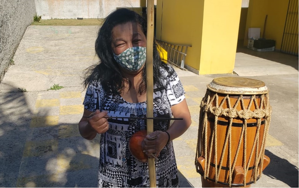 Senhora tocando os instrumentos utilizados durante as aulas de Maculelê. Na foto, ela toca berimbau com um atabaque ao lado.