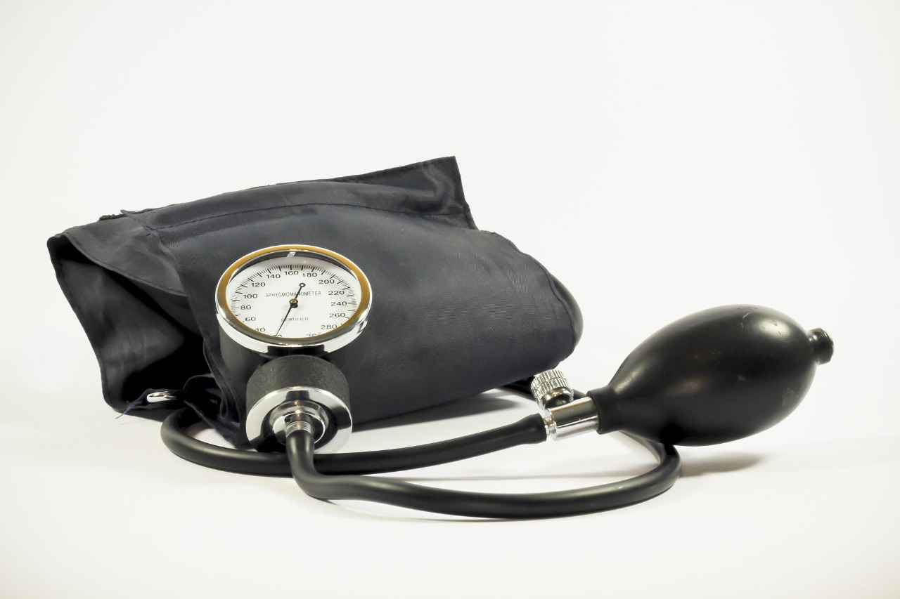 Em fundo branco há um medidor de pressão arterial na cor preta com relógio e bombinha para apertar.