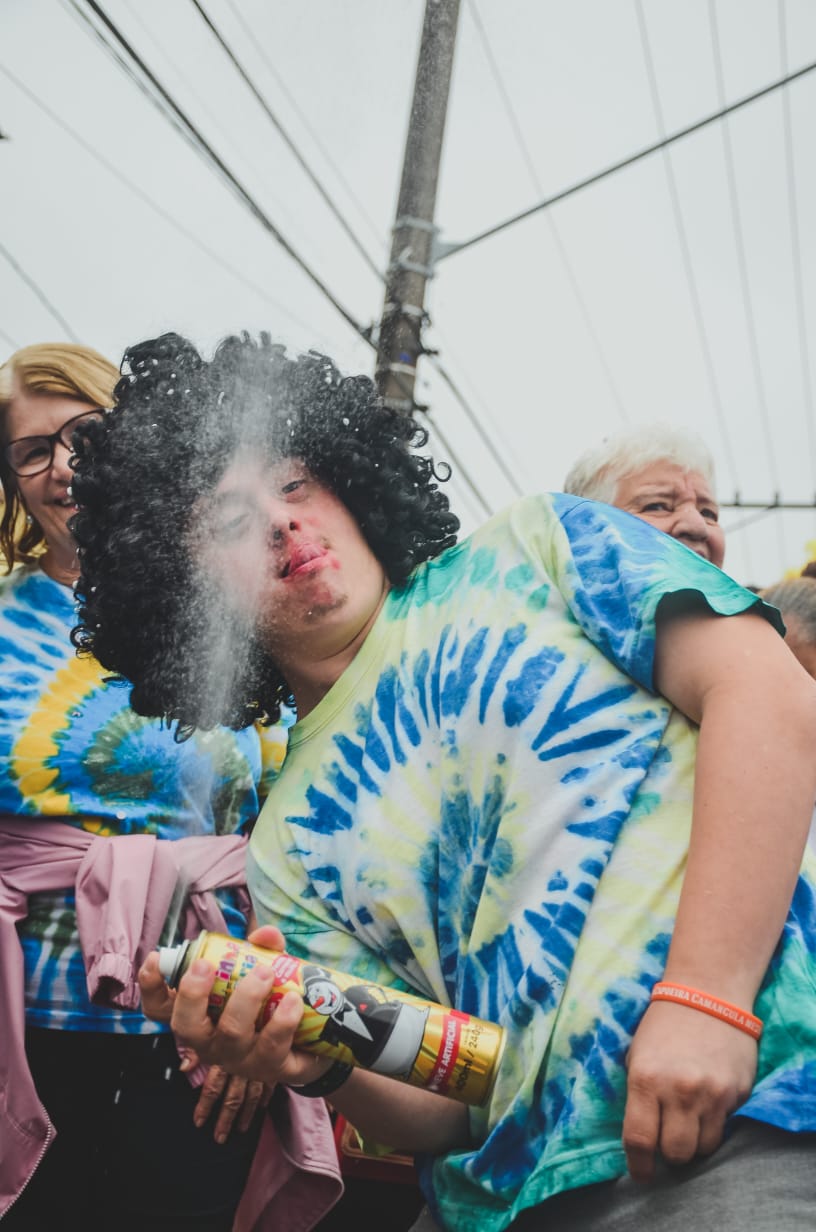 Um homem com Síndrome de Down está brincando com um spray. Ele usa camiseta colorida azul e verde, e usa uma peruca com cabelos cacheados enormes.