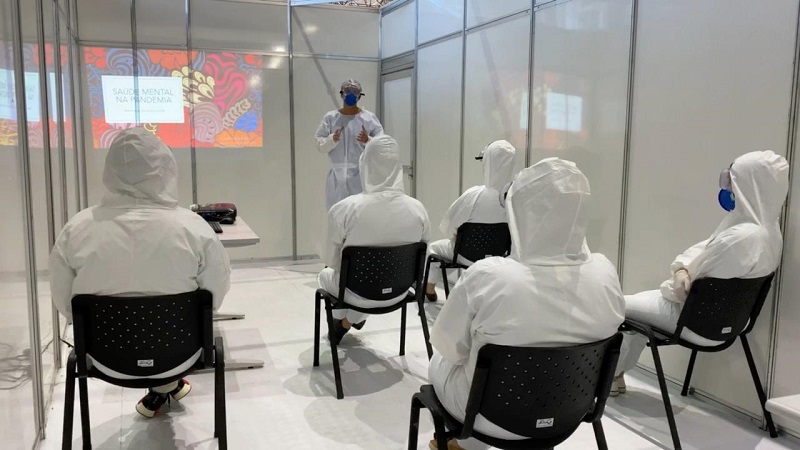 #PraCegoVer: Uma sala com cinco pessoas sentadas em cadeiras separadas. Eles estão ouvindo instruções de um outro homem, sobre saúde mental na pandemia. Há uma apresentação com o tema 