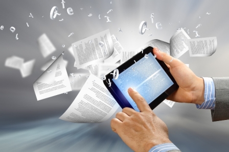 No centro da foto, um tablet é utilizado por uma pessoa para manusear um documento enquanto ao redor do tablet diversas folhas de papel estão voando/sendo jogadas ao alto. 