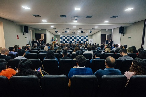Foto de auditório com pessoas sentadas em cadeiras, assistindo uma coletiva de imprensa