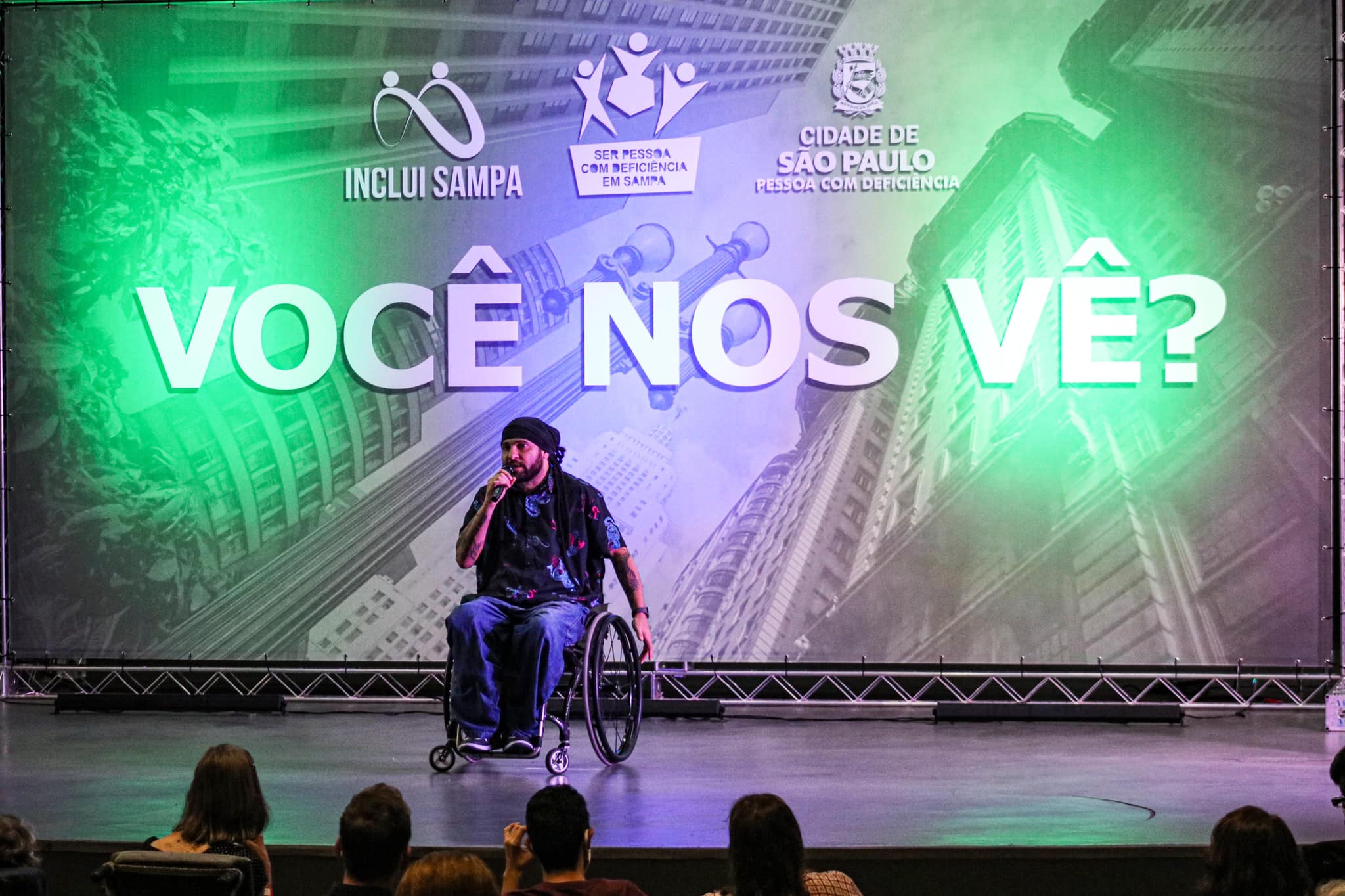 no palco do evento, o músico e ativista Billy Saga, que fala ao microfone. Ele usa uma cadeira de rodas.