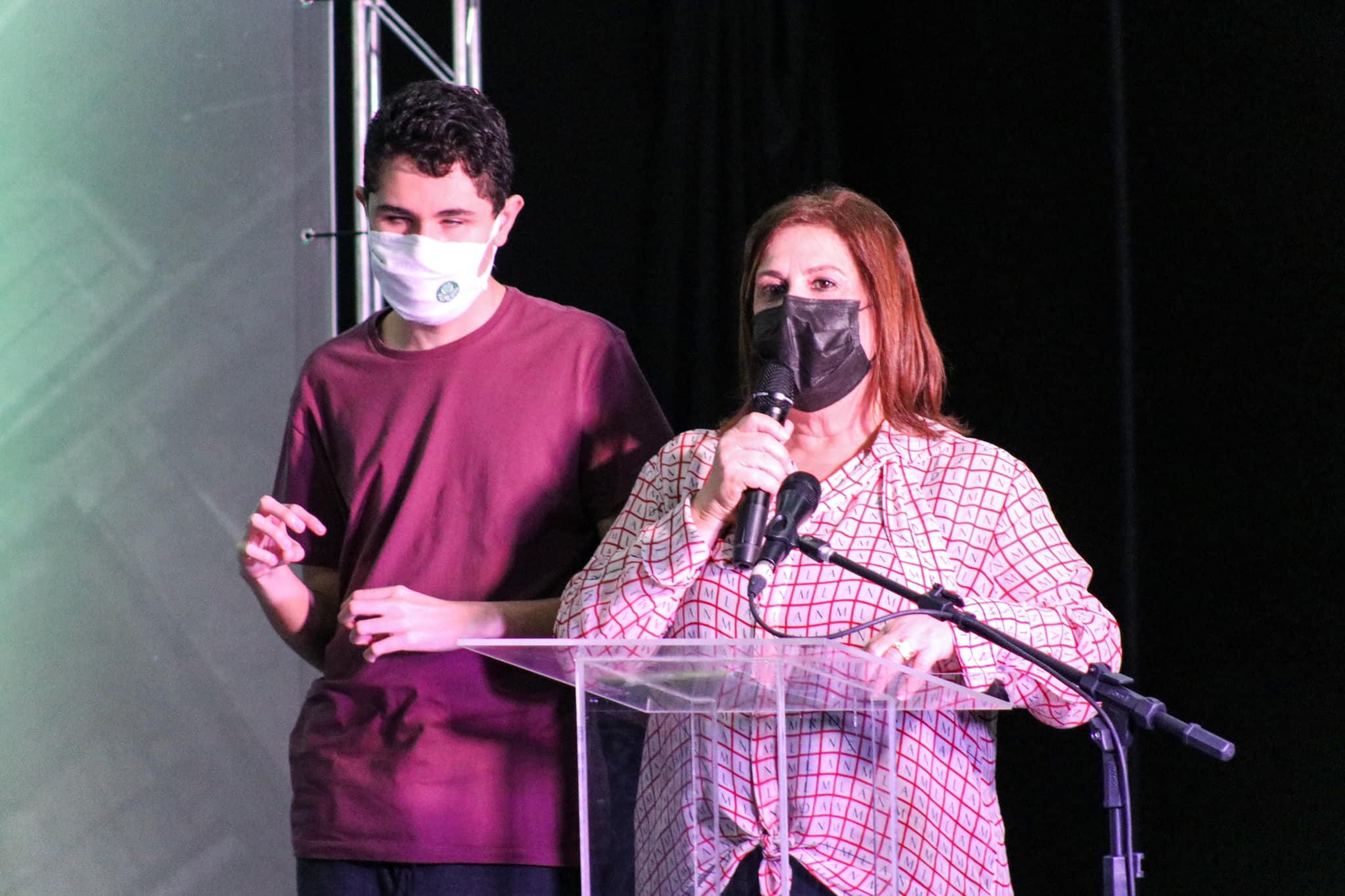 no palco do evento, a secretária da SMPED Sílvia Grecco ao lado do seu filho Nichollas Grecco. Ela fala ao microfone.