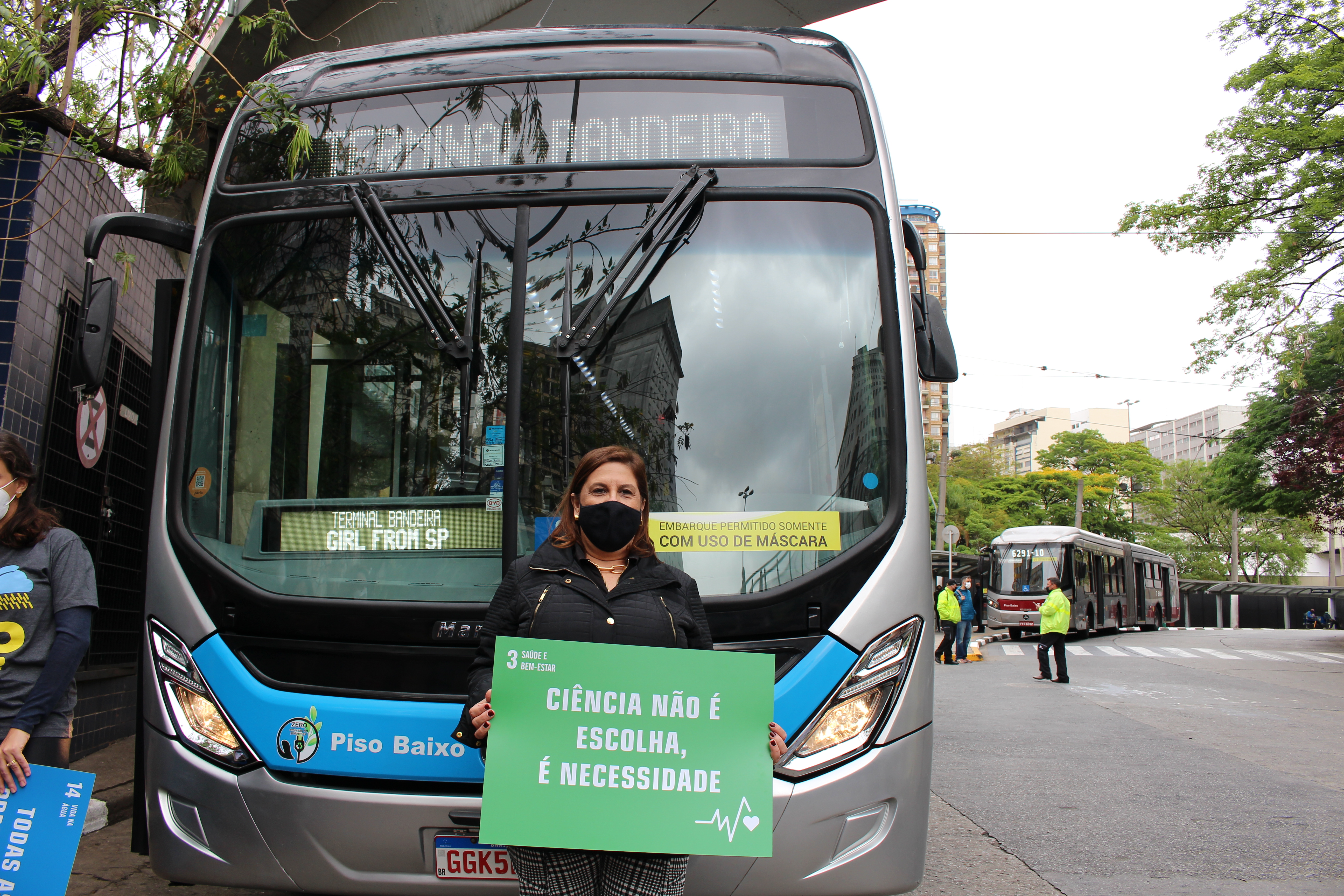 A secretária Silvia Grecco está na frente de um ônibus segurando um cartaz na cor verde com o texto: Saúde e Bem-Estar. ''Ciência não é escolha, é necessidade''