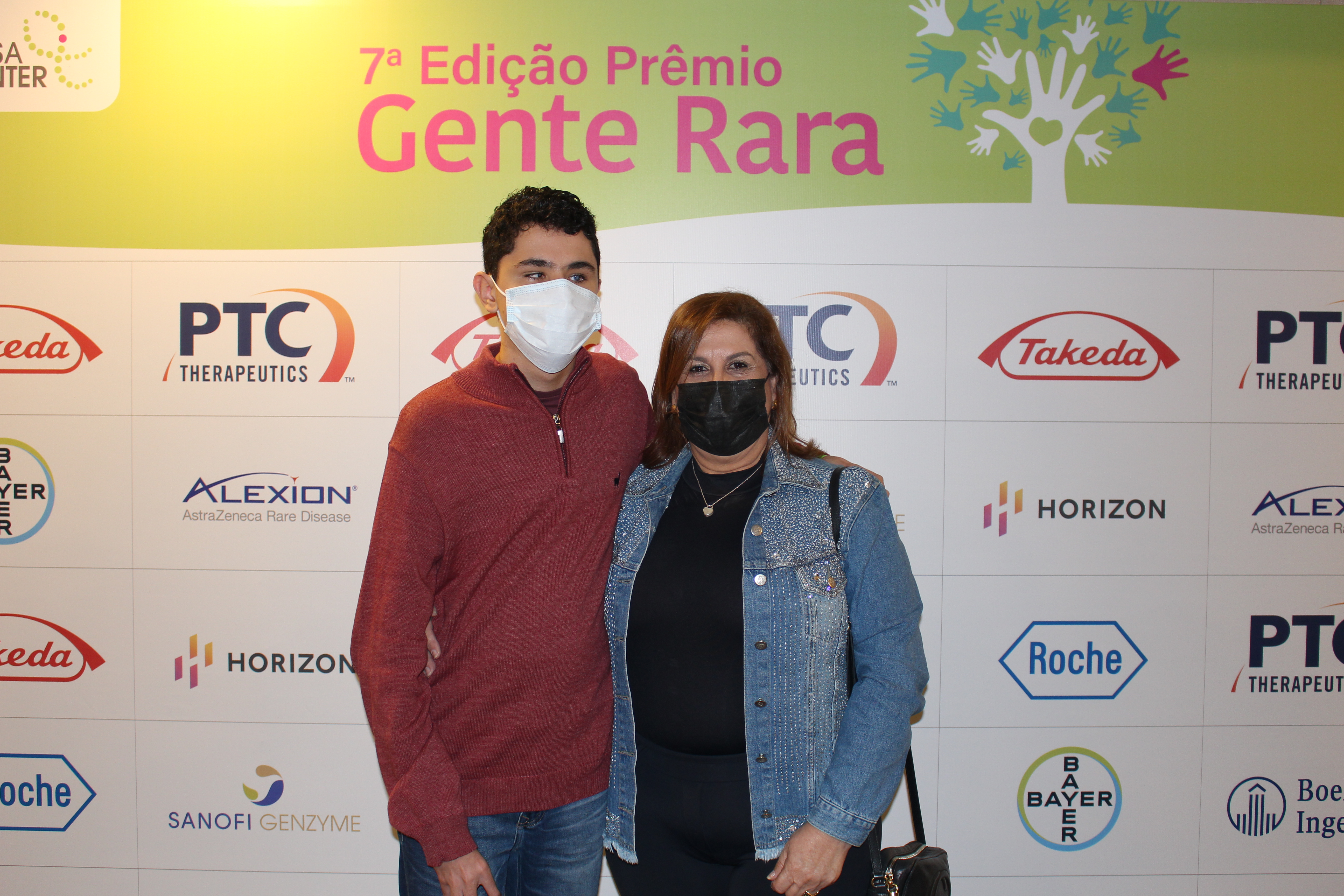 A secretária Silvia Grecco e seu filho Nickollas Grecco estão na frente do backdrop do evento. Nele contém os logotipos de empresas que apoiam o evento.