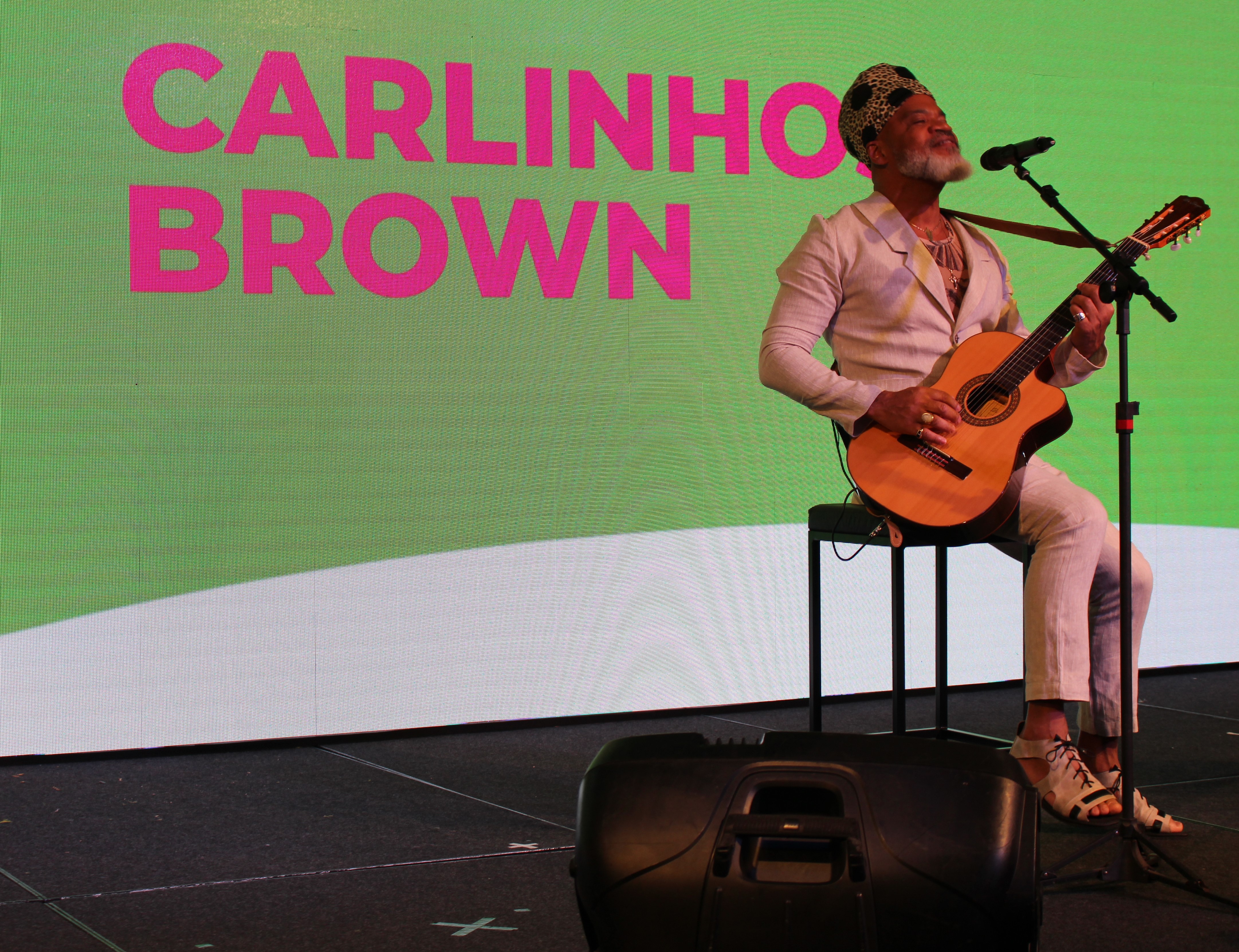 Carlinhos Brown está no palco, sentado em um banco, segurando um violão e com um microfone á sua frente. Ele canta para o público e atrás dele o telão está nas cores verde e branca com o texto na cor rosa: Carlinhos Brown.